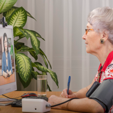 Older white woman, sitting at desk with blood pressure cuff on and looking at telehealth appointment on computer