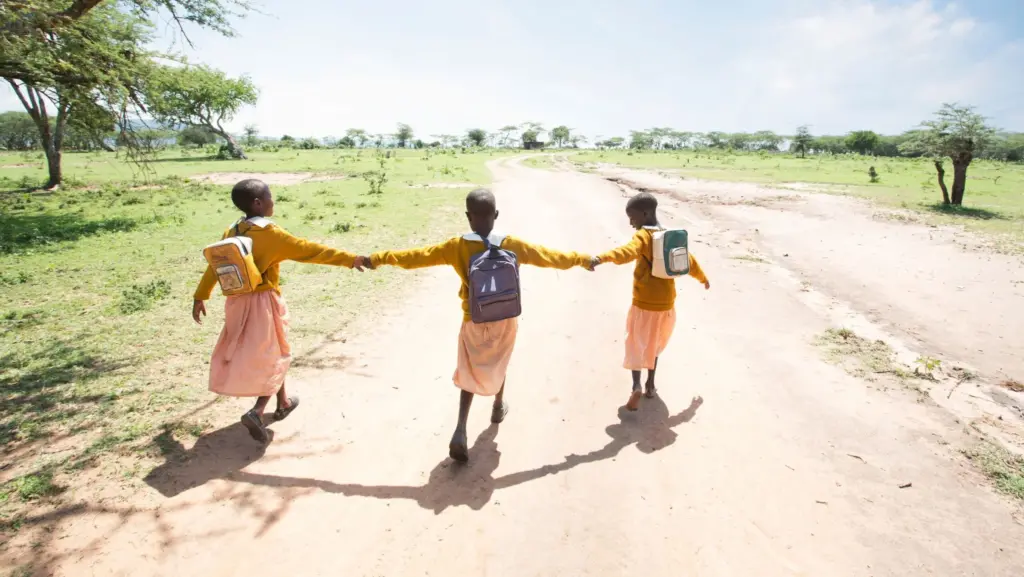 three girls in yellow sweaters and pink skirts holding hands walking down a dirt road